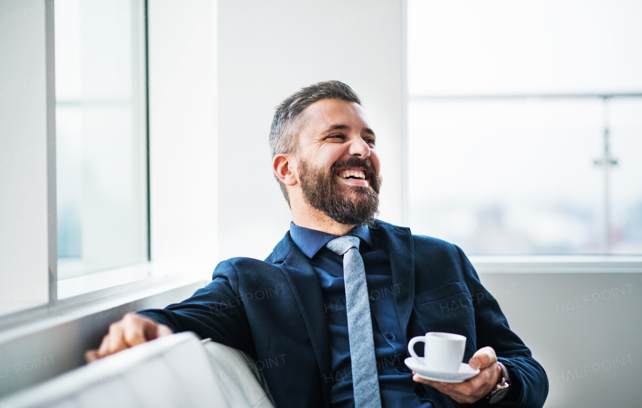 A portrait of businessman with a cup of coffee sitting on a white sofa in a modern office, laughing. Copy space.