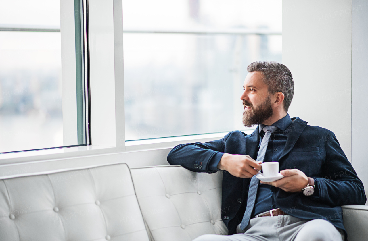 A portrait of businessman with a cup of coffee sitting on a white sofa in a modern office, looking out of a window. Copy space.