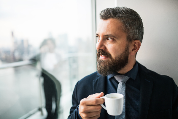 A close-up portrait of businessman with a cup of coffee looking out of a window in an office.