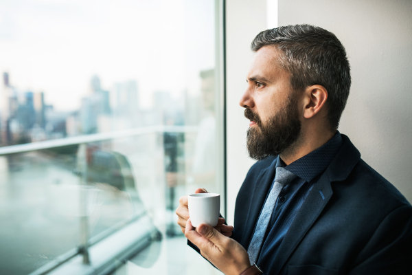 A close-up portrait of businessman with a cup of coffee looking out of a window in an office.