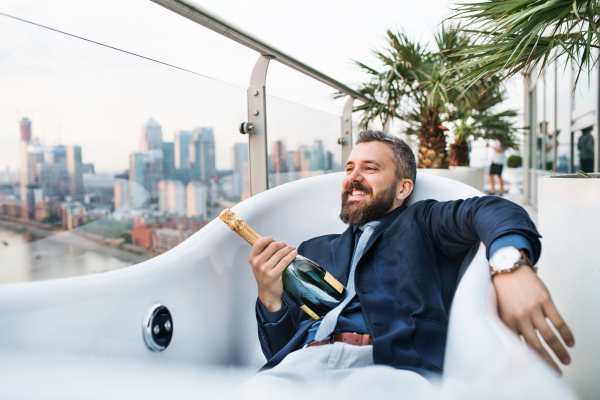 Businessman with a bottle of champagne lying in empty hot tub, London view panorama in the background. Copy space.