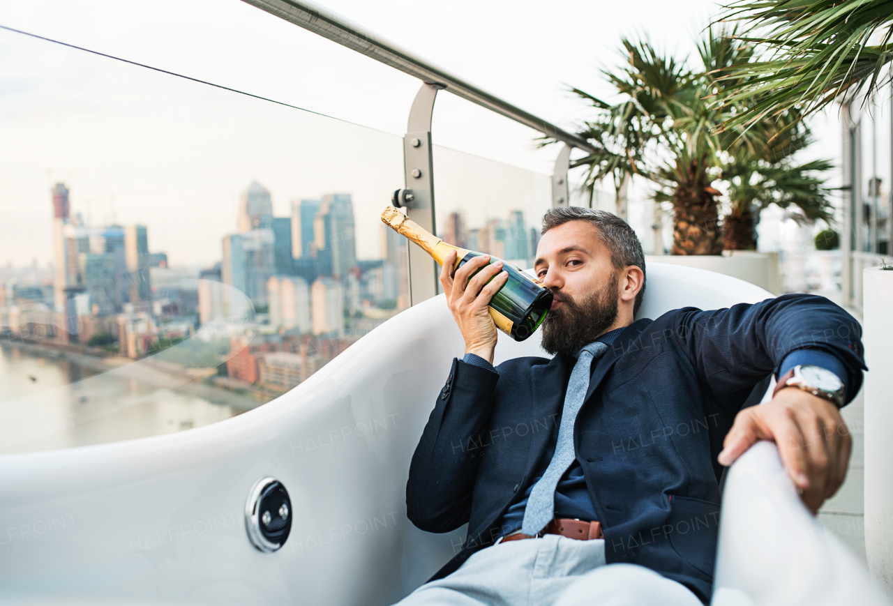 Businessman with a bottle of champagne lying in empty hot tub, London view panorama in the background. Copy space.