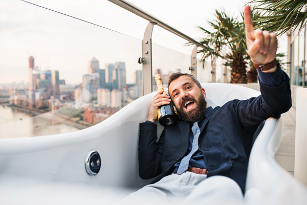 Businessman with a bottle of champagne lying in empty hot tub, London view panorama in the background. Copy space.