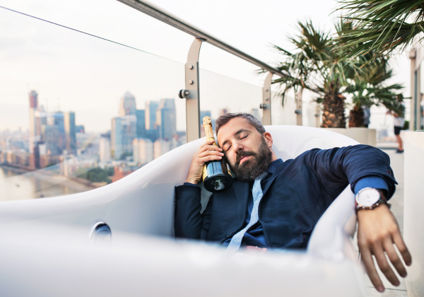 Businessman with a bottle of champagne lying in empty hot tub, London view panorama in the background. Copy space.