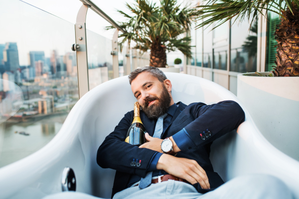 Businessman with a bottle of champagne lying in empty hot tub, London view panorama in the background. Copy space.