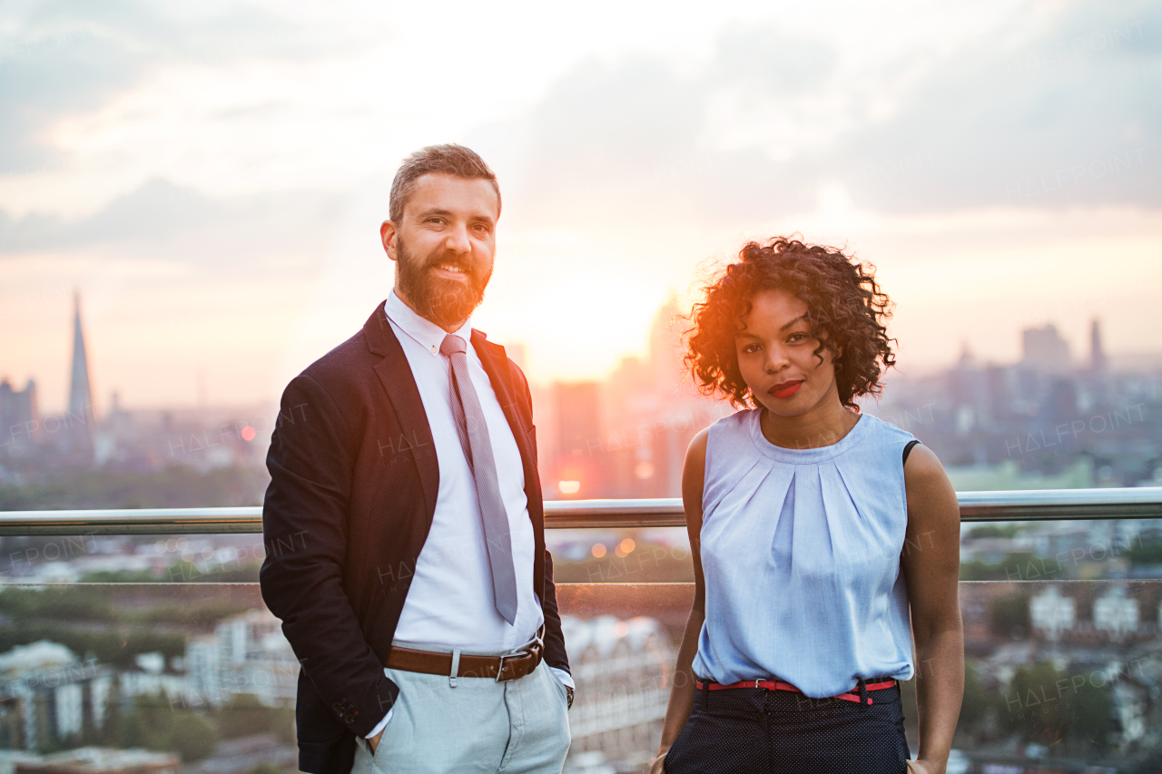 A portrait of businesspeople standing against London rooftop view at sunset, hands in pockets.