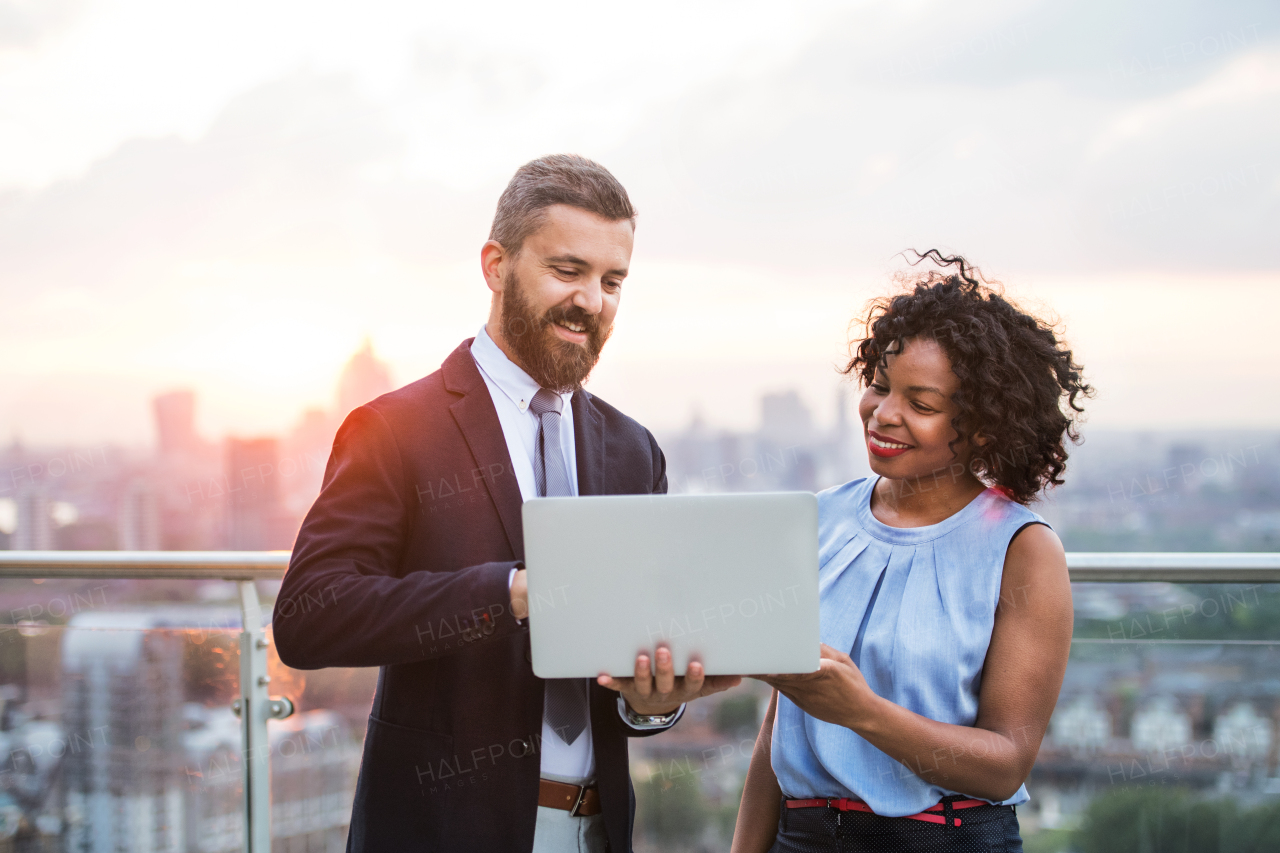 A portrait of businesspeople with laptop standing against London sunset view, talking.