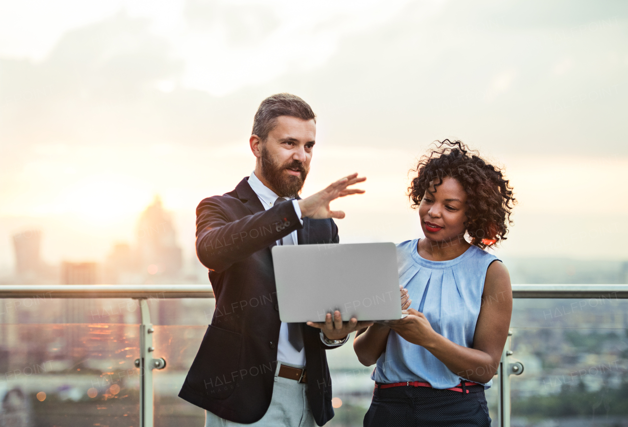 A portrait of businesspeople with laptop standing against London sunset view, talking.