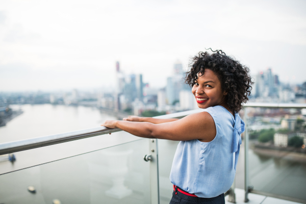 A portrait of a laughing black woman standing on a terrace in London, holding railing. Copy space.