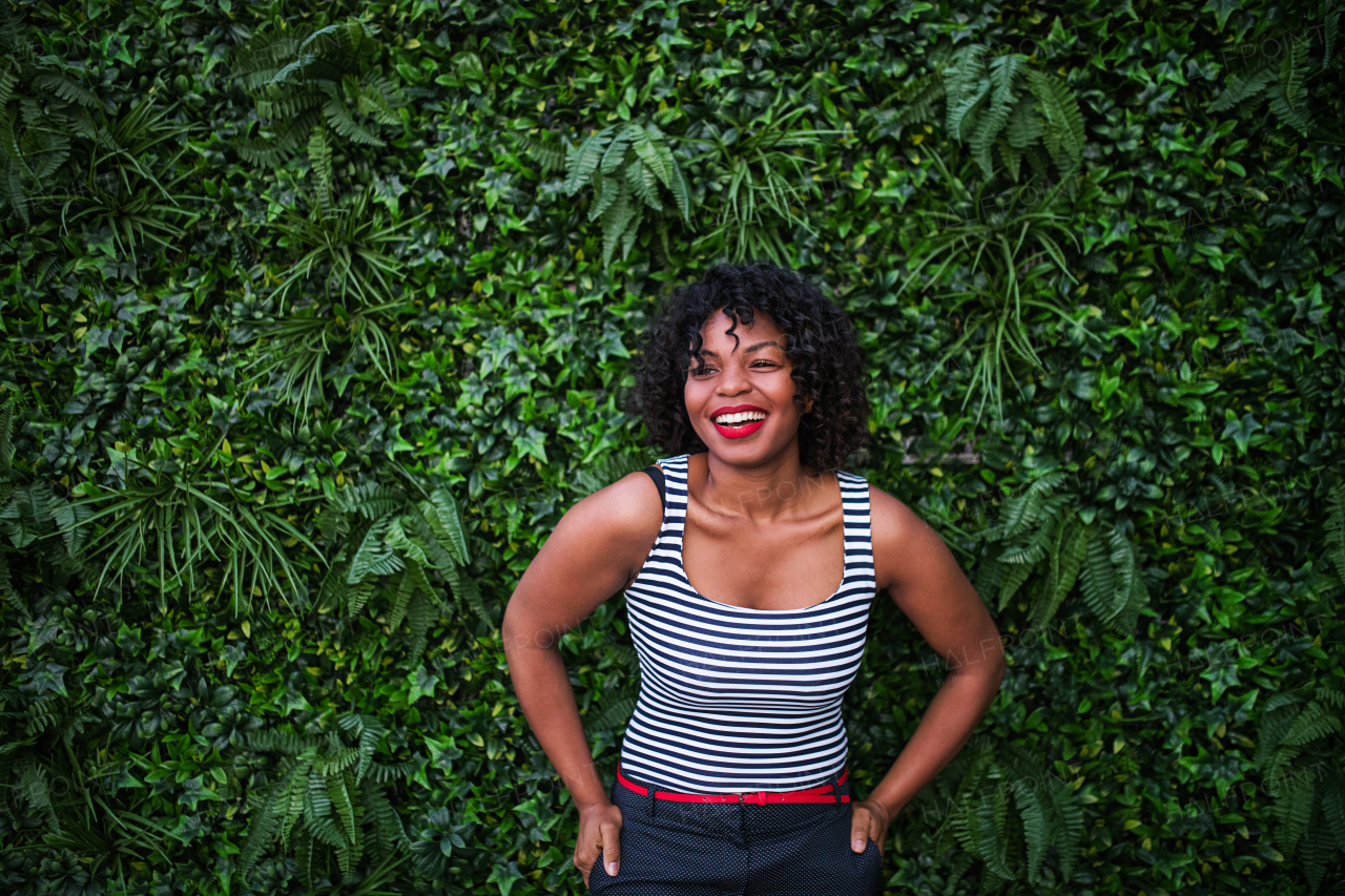 A portrait of a black laughing woman standing against green background of bush leaves.