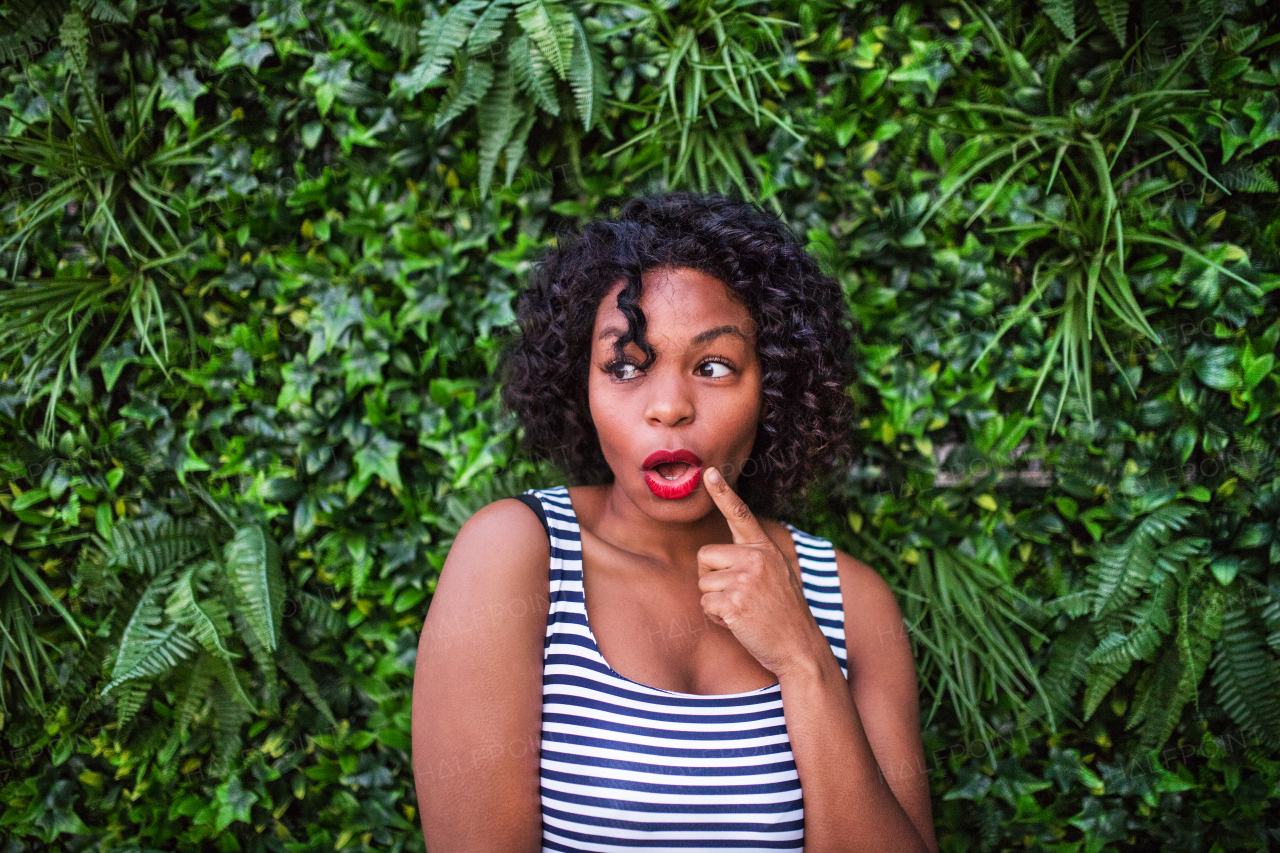 A portrait of a suprised woman standing against green background of bush leaves.