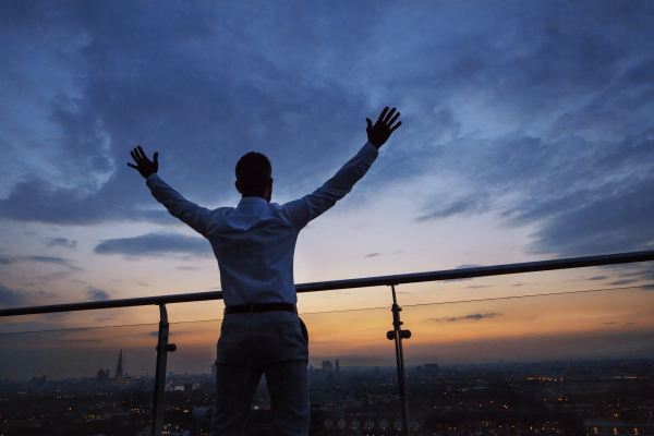 A rear view of hipster businessman standing against London view panorama at dusk, raising hands. Copy space.