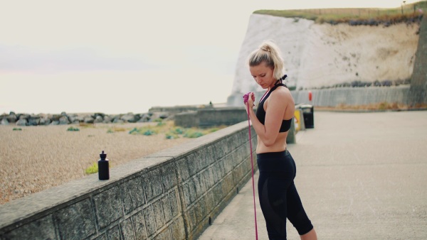 Young sporty woman runner doing exercise with elastic rubber bands outside on a beach in nature.