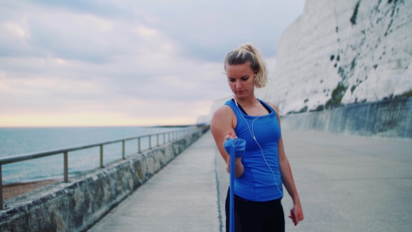 Young sporty woman runner with earphones doing exercise with elastic rubber bands outside on a beach in nature.