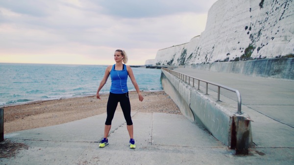 Young sporty woman runner with earphones stretching outside on the beach, listening to music.