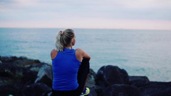 A rear view of young sporty woman runner sitting on rocks on the beach outside.