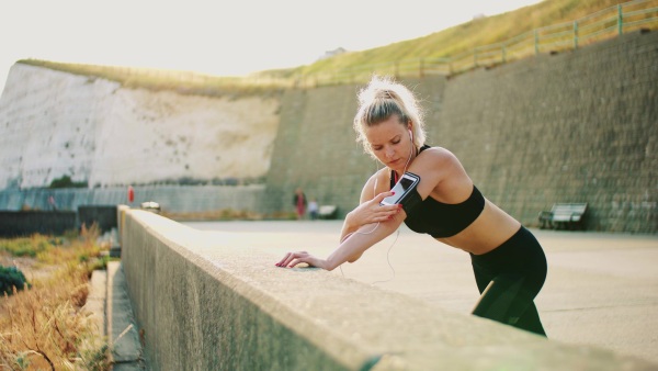 Young sporty woman runner with earphones and smartphone in armband stretching outside on the beach in nature, listening to music.