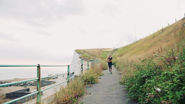 Young sporty woman runner with smartphone in armband running outside on the beach in nature. Slow motion.