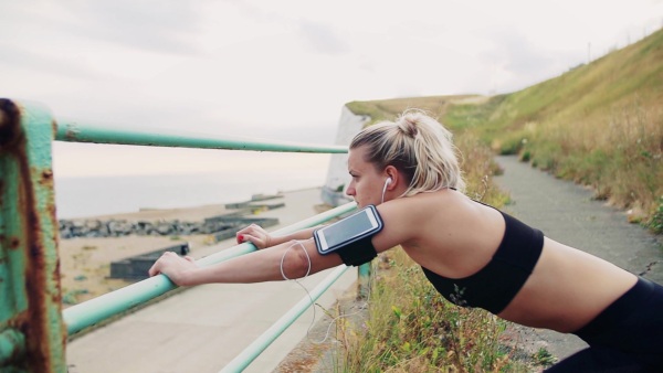 Young sporty woman runner with earphones and smartphone in armband stretching outside on the beach in nature, listening to music. Slow motion.