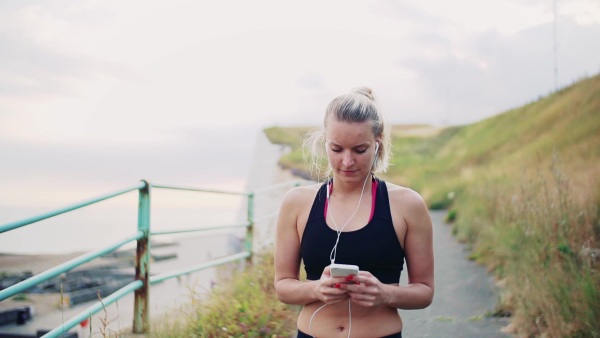 Young sporty woman runner walking outside on the beach in nature, using smartphone. Slow motion.