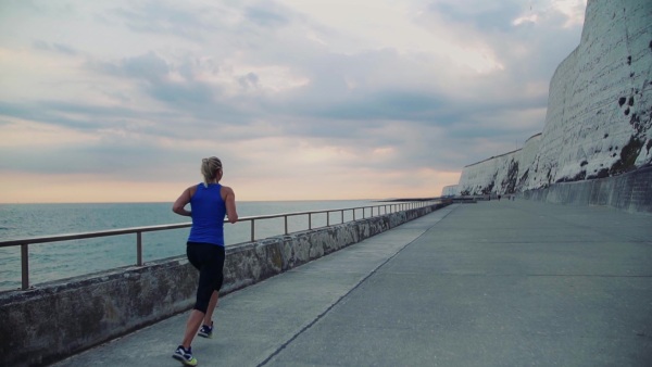 Young sporty woman runner with earphones running by the sea outside in nature, listening to music. Rear view. Slow motion.