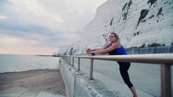 Young sporty woman runner with earphones stretching on a railing outside on the beach, listening to music. Slow motion.