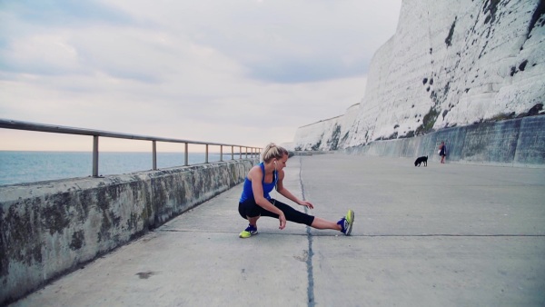 Young sporty woman runner with earphones stretching outside on the beach, listening to music. Slow motion.