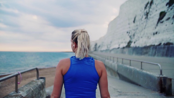 Rear view of a young sporty woman runner with earphones walking outside on the beach in nature, listening to music and looking back. Copy space. Slow motion.