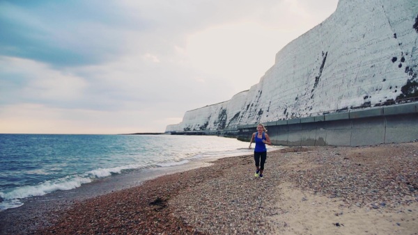 Young sporty woman runner with earphones in blue sportswear running outside on the beach in nature. Copy space. Slow motion.