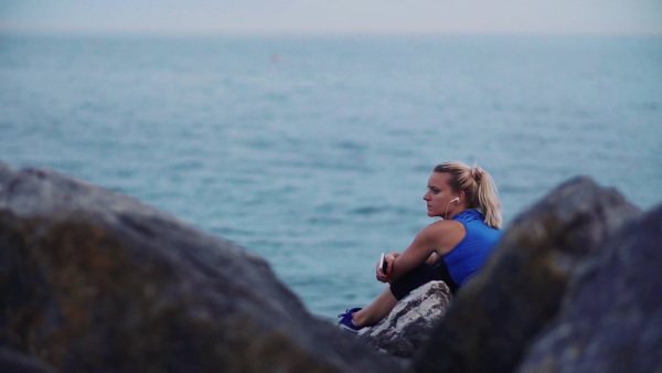 Young sporty woman runner with earphones sitting on rocks on the beach outside, listening to music. Slow motion.