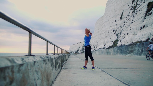 Young sporty woman runner with earphones stretching outside on the beach, listening to music.