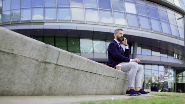 Hipster businessman with coffee and smartphone sitting on a bench on the street in London, making a phone call.