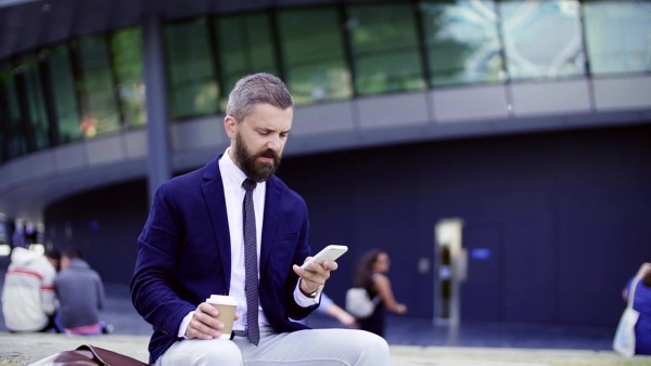 Hipster businessman with smartphone sitting on a bench on the street in London, text messaging and drinking coffee. Slow motion.