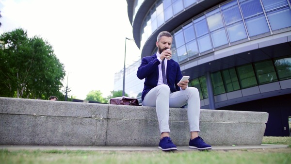 Hipster businessman with coffee and smartphone sitting on a bench on the street in London, text messaging. Slow motion.