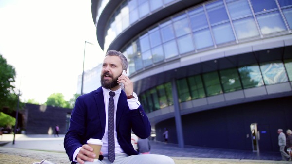 Hipster businessman with coffee and smartphone sitting on a bench on the street in London, making a phone call. Slow motion.