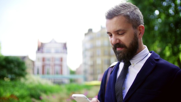 Hipster businessman with smartphone standing on the street in city, text messaging. Slow motion.