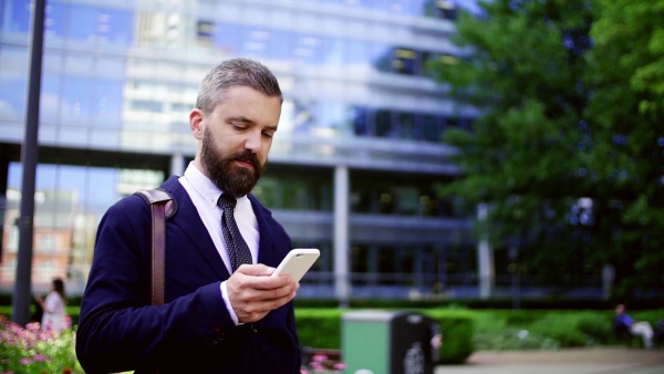 Hipster businessman with smartphone standing on the street in city, text messaging. Slow motion.