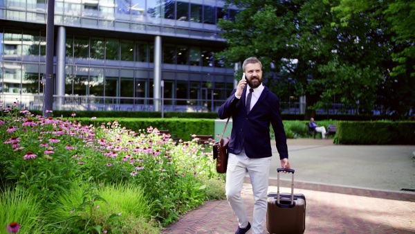 Hipster businessman with laptop bag, suitcase and smartphone walking in park in London, making a phone call. Slow motion.