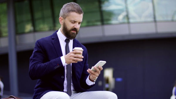 Hipster businessman with coffee and smartphone sitting on a bench on the street in London, text messaging.