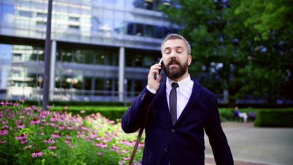 Hipster businessman with laptop bag and smartphone walking in park in London, making a phone call. Slow motion.