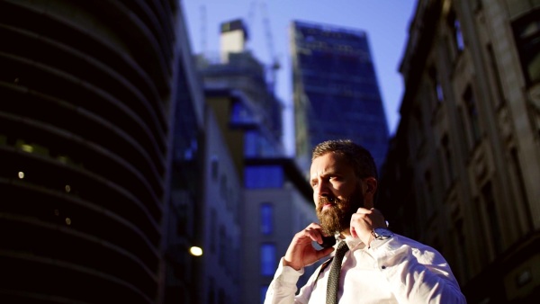 Hipster businessman standing on the street in London in the evening, putting headphones on head and listening to music. Copy space. Slow motion.