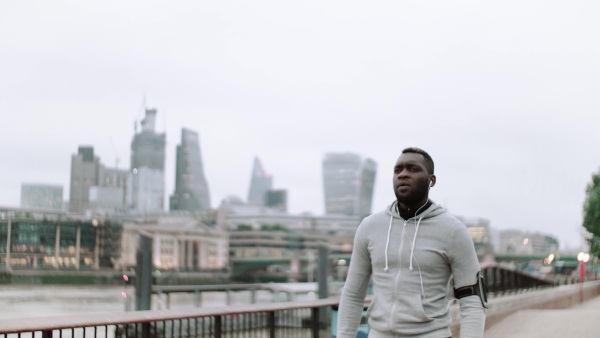 Young sporty black man runner with smartwatch, earphones and smart phone in arm band running on the bridge outside in a city.