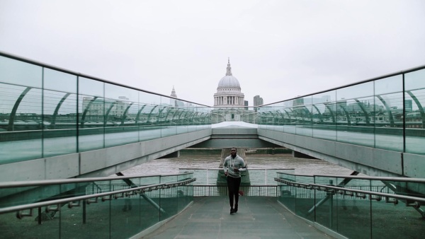 Young sporty black man runner with smart phone in arm band running on the bridge outside in a London city, St. Paul's cathedral in the background. Slow motion.