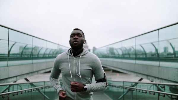 Young sporty black man runner with smart phone in arm band running on the bridge outside in a London city, St. Paul's cathedral in the background. Slow motion.