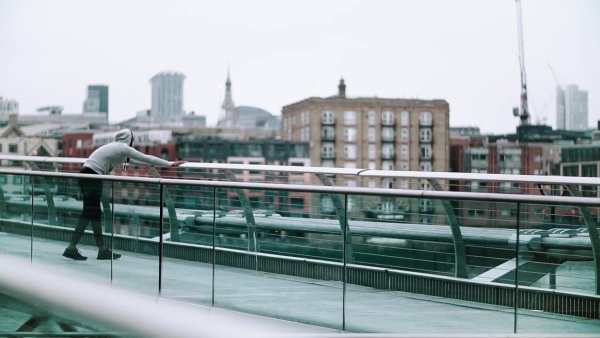 Young sporty black man runner on the bridge in a city, stretching. Slow motion.