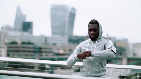 Young sporty black man runner with smartwatch, earphones and smartphone in an armband on the bridge in a city, resting. Slow motion.