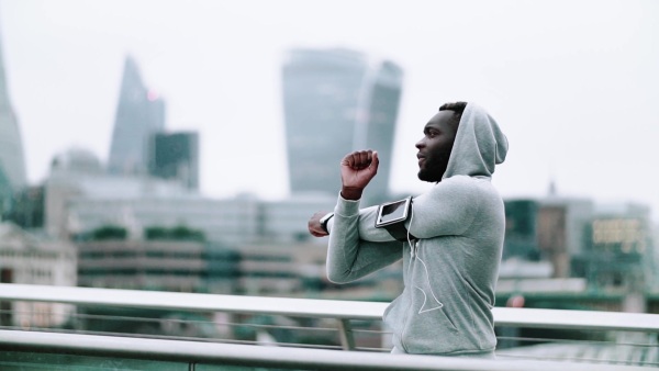 Young sporty black man runner with smartwatch, earphones and smartphone in an armband on the bridge in a city, stretching. Slow motion.