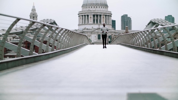 Young sporty black man runner with smart phone in arm band running on the bridge outside in a London city, St. Paul's cathedral in the background. Slow motion.