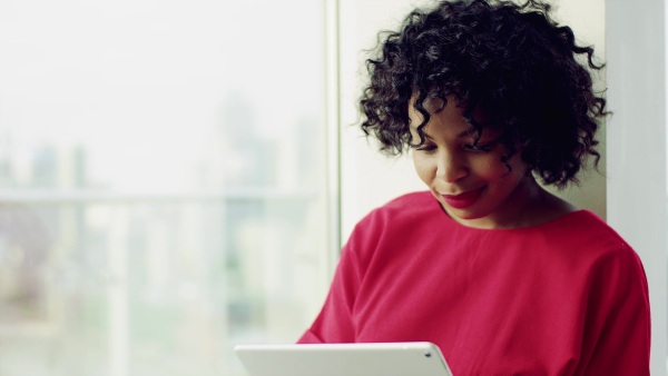 A portrait of woman standing by the window against London rooftop view panorama, using tablet. Copy space.