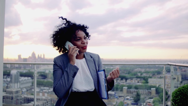 A portrait of black businesswoman standing on a terrace against London rooftop view panorama, making a phone call. Copy space.